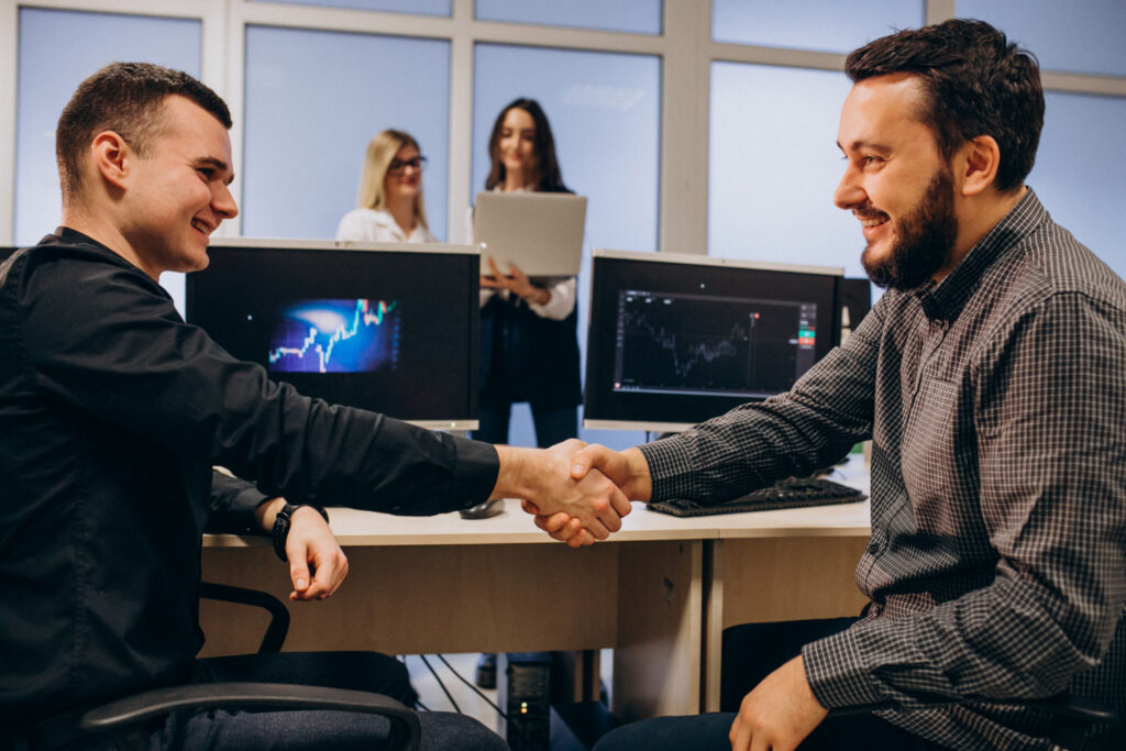 Dois homens sentados em uma mesa de trabalho, de frente um para o outro apertando as mãos