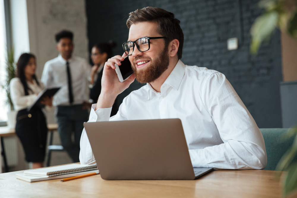 Um homem sentado em frente ao notebook conversando alegremente no telefone. Duas pessoas ao fundo desfocadas